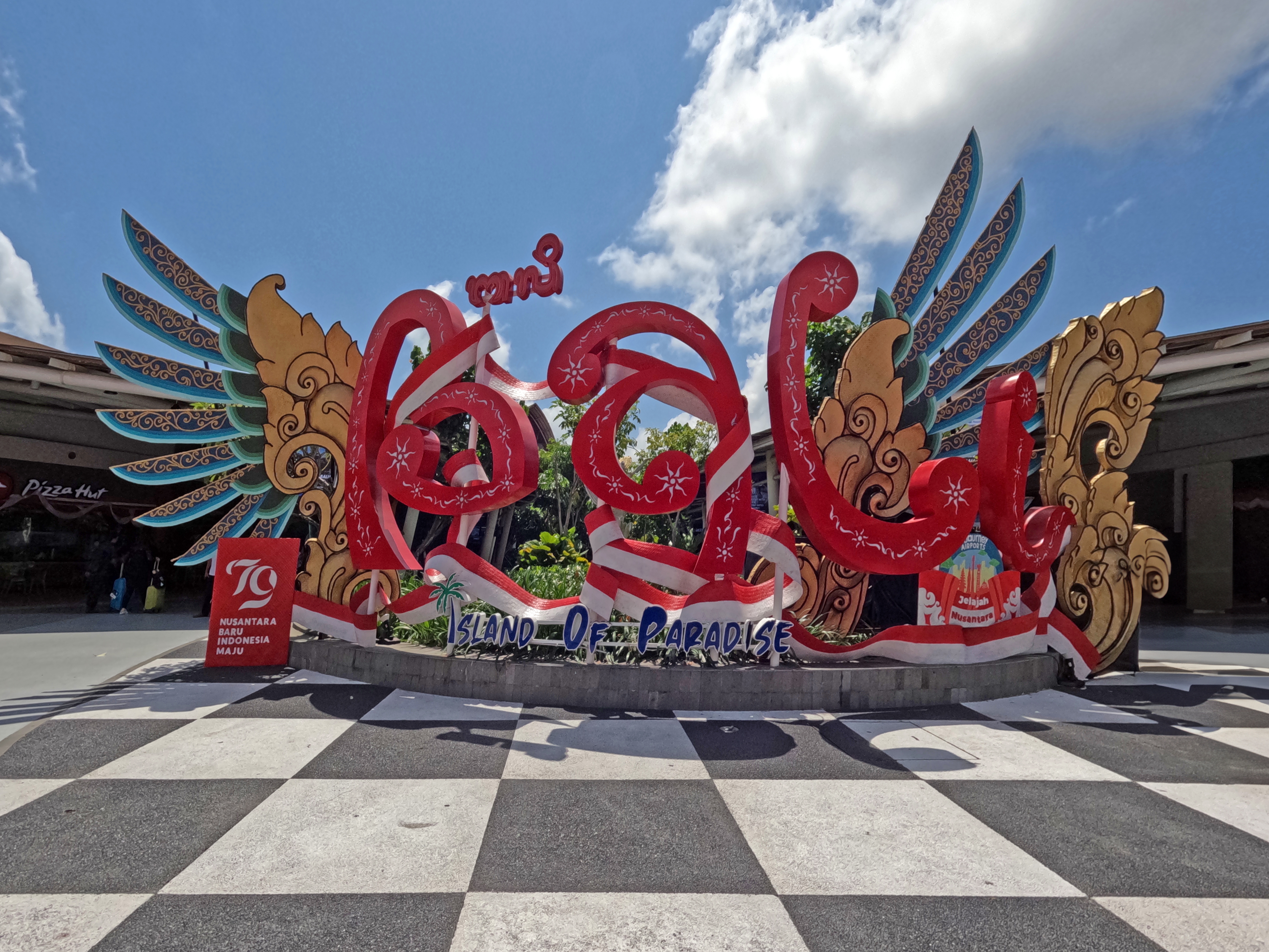 Colourful &ldquo;Bali &ndash; Island of Paradise&rdquo; sign framed by ornate winged carvings under a bright blue sky, marking a lively public space in Bali and setting the scene for travel in Canggu.